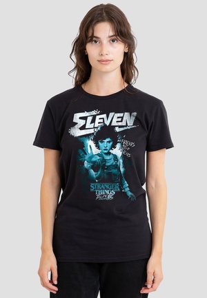 Young woman wearing black Stranger Things Eleven graphic tee with character raising a hand and text "Friends Help Friends."