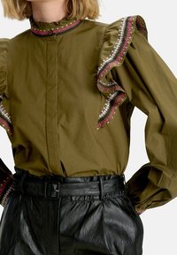 Blusa verde oliva con maniche a palloncino, scollatura arricciata e accenti ricamati colorati con cuciture rosa, bianche e nere. Pantaloni in pelle nera.