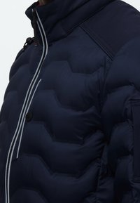 Ναυτική μπουφάν με πλούσιο γεωμετρικό σχέδιο, έντονες οριζόντιες ραφές, κούμπωμα με κουμπιά και αντίθετα μανίκια σε διαφορετική απόχρωση.