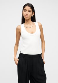 Mujer con cabello largo y oscuro, vestida con una blusa blanca sin mangas con escote redondo, pantalones negros de cintura alta, collar de perlas y pendientes de aro plateados.