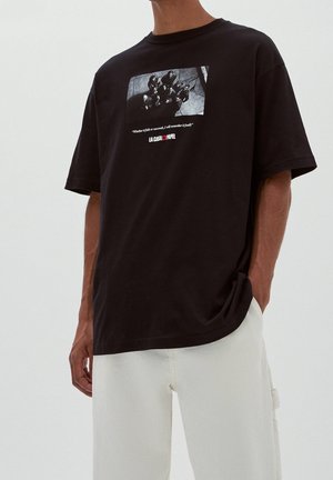 Mand iført en sort T-shirt med et sort-hvidt gruppefoto og tekst fra "La Casa de Papel" på brystet, kombineret med hvide bukser.