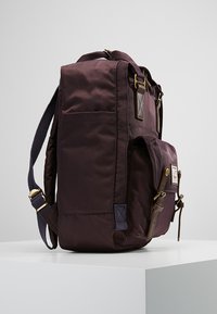Dark purple nylon backpack with adjustable straps, a front pocket, and gold hardware accents. Features a rectangular shape and textured surface.