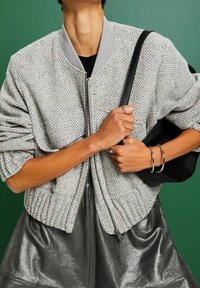 Personne portant une veste zippée grise texturée et une jupe noire brillante, tenant un sac à bandoulière noir avec des bracelets et des bagues argentés sur un fond vert.