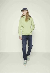 Ανοιχτό πράσινο φούτερ με μπροστινή τσέπη, σκούρο μπλε τζιν και πολύχρωπα αθλητικά παπούτσια. Φοράει μπλε καπέλο και στέκεται σε ουδέτερο φόντο.