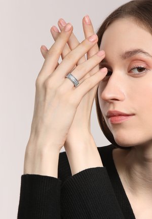 Silberring mit strukturiertem Oberflächen, ovale Form, am Zeigefinger getragen. Das Modell hat helle Haut und kurze, helle Nägel, die Hände sind entspannt mit gekreuzten Fingern.