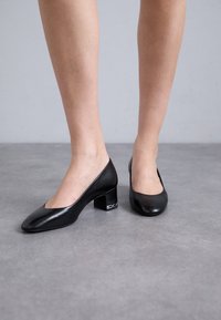 Sorte skinnpumps med lav blokkhelt og åpen, rund tå. Har subtil søm og en liten metallisk detalj på helen.