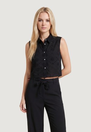 Donna bionda che indossa un crop top nero senza maniche con bottoni e ritagli floreali e pantaloni neri a vita alta con cintura a fiocco.