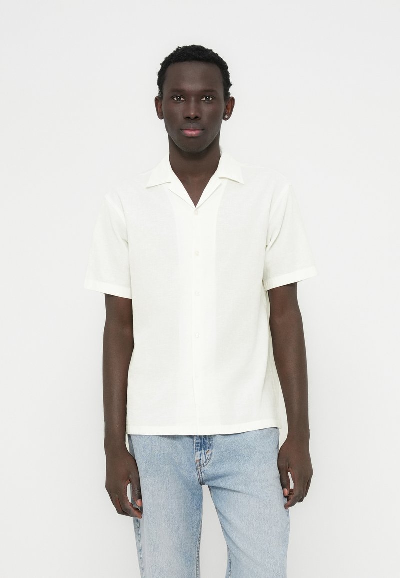 Πουκάμισο με κοντά μανίκια και γιακά από ελαφρύ εκρού ύφασμα, με κουμπιά στο μπροστινό μέρος και υφή φινιρίσματος. Φορεμένο με ανοιχτόχρωμο μπλε τζιν παντελόνι.