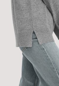 Grauer Strickpullover mit geripptem Saum und seitlichen Schlitzen, kombiniert mit hellblauen Jeans im klassischen geraden Schnitt und sichtbaren Nähten.
