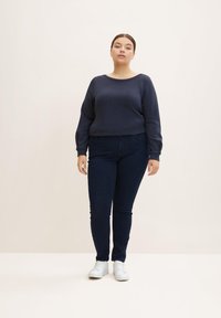 Sudadera azul marino con cuello redondo, llevada con jeans de mezclilla oscuros. El modelo usa zapatillas blancas y está de pie sobre un fondo claro.