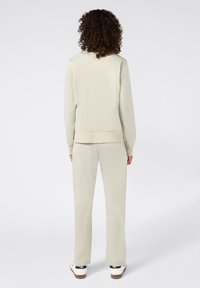 Tuta beige con un design attillato e polsini a coste. Texture liscia. I pantaloni hanno una vestibilità dritta, indossati con sneakers bianche.