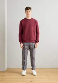 Sudadera burdeos con cuello redondo y pequeño logo, combinada con pantalones gris a rayas y zapatillas blancas sobre un suelo de madera.