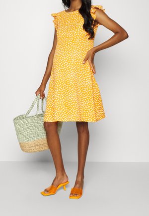 Żółta sukienka w kropki z falbaniastymi rękawami i rozkloszowanym dołem. W połączeniu z jasnozieloną torbą z naturalnego włókna oraz pomarańczowymi sandałami na obcasie.