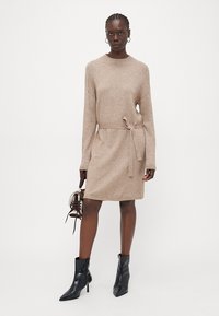 Abito in maglia beige con maniche lunghe, scollo rotondo e cintura in vita. Indossato con stivaletti neri alla caviglia e una piccola borsa a tracolla. Texture liscia.