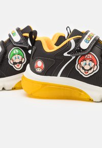 Zwarte sneakers met oranje binnenkant en gele doorzichtige zool. Heeft patches van de personages Mario en Luigi, witte accenten en klittenbandsluitingen.