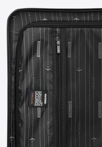 Intérieur de valise noire présentant une doublure texturée avec des motifs d'avion blancs et de lignes pointillées, ainsi qu'une poche en mesh avec une fermeture éclair.