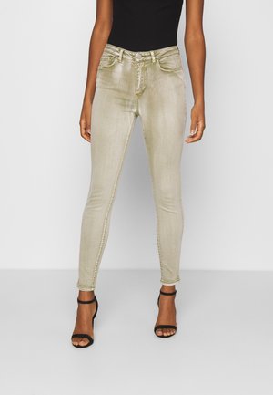 Kobieta w jasnobeżowych obcisłych jeansach i czarnych sandałach na wysokim obcasie z paskami na tle jednolitej białej ściany.