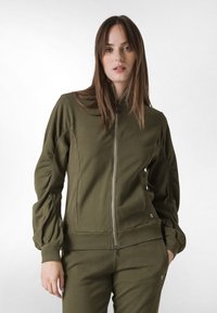 Giacca verde oliva con zip realizzata in tessuto morbido, con maniche a sbuffo e vita aderente, abbinata a pantaloni coordinati.