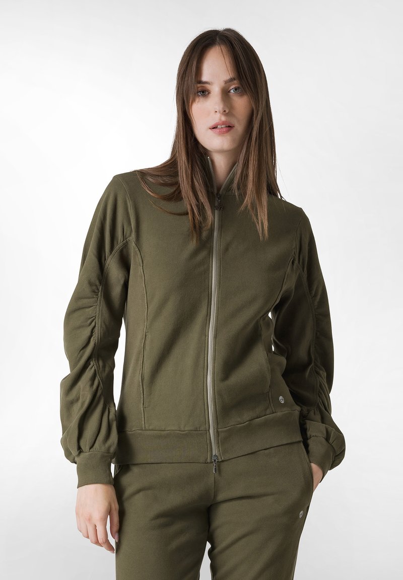 Giacca verde oliva con zip realizzata in tessuto morbido, con maniche a sbuffo e vita aderente, abbinata a pantaloni coordinati.