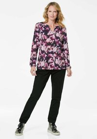 Lila- und rosa-blumengemusterte Langarmbluse mit V-Ausschnitt; kombiniert mit schwarzen Hosen und silbernen, hochgeschnittenen Sneakern.