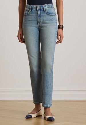 Naine, kes kannab helerohelisi kõrge vöökohaga teksaseid, musta pluusi, kilpkonnakujulist käevõru ning kahetoonilisi beeže ja mereväe balletikingi, seisab puidust põrandal.