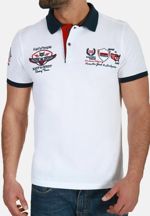 Mand iført hvid polo-shirt med sort krave og røde accenter, med raceteam-logoer og broderet tekst på fronten og ærmerne.