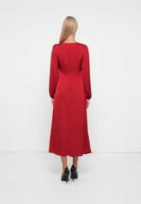 Robe mi-longue rouge en satin avec des manches longues, un dos carré décolleté et un corsage ajusté, associée à des chaussures à talons hauts noires.