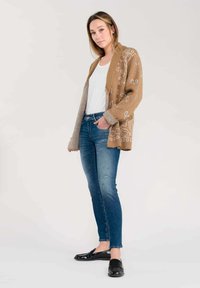 Cardigan lavorato a maglia marrone con motivi bianchi, indossato sopra una camicia bianca e jeans blu. Il cardigan ha una vestibilità comoda e polsini a coste.