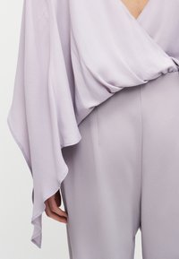 Μπλούζα σε λιλά με φαρδιές ροές μανικιών σε συνδυασμό με ψηλόμεσο, ριγμένο παντελόνι, εμφανίζοντας το χέρι που ακουμπάει στο πλάι.