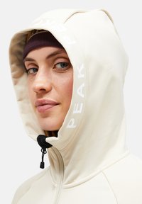 Cream-colored hooded jacket with a black drawcord, logo printed on the hood, and a smooth texture. Zipper closure is visible on the front.