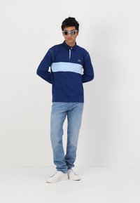 Mornarska polo majica z dolgimi rokavi s svetlo modro horizontalno črto in logotipom "LACOSTE". V kombinaciji s svetlo modrimi kavbojkami in belimi športnimi copati.