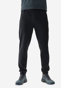 Pantalones deportivos negros con cintura el�ástica, diseño de pierna acampanada y bolsillos laterales con cremallera, combinados con zapatillas de correr grises.