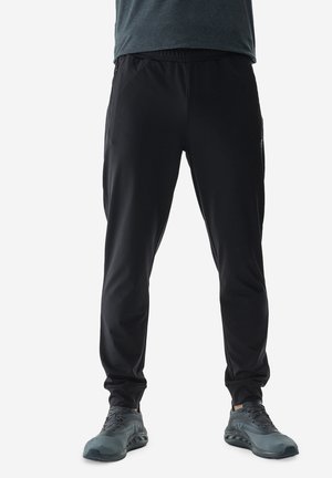 Pantalones deportivos negros con cintura elástica, diseño de pierna acampanada y bolsillos laterales con cremallera, combinados con zapatillas de correr grises.