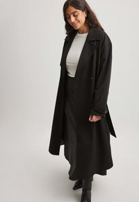 Sort trenchcoat med dobbeltknapning, brede revers og lange ærmer. Bæres over en tætsiddende hvid top, parret med en lang sort nederdel.