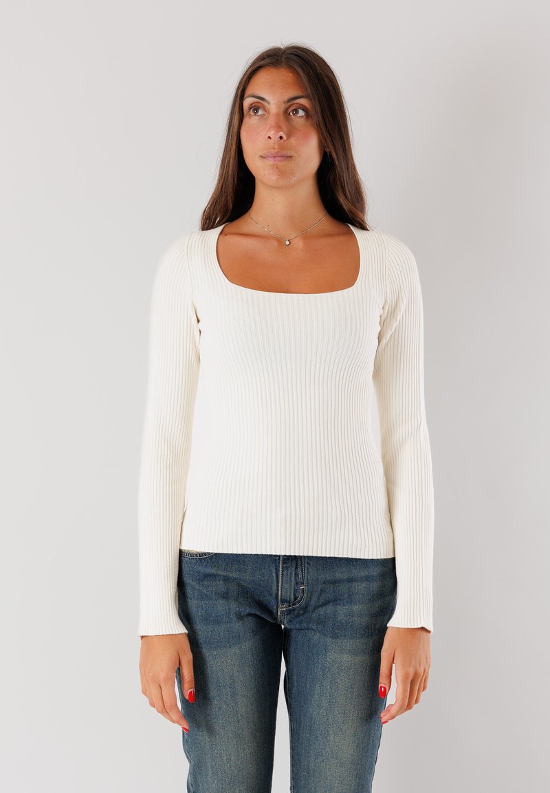 Top lungo a maniche lunghe bianco a costine con scollatura quadrata, forma aderente e texture morbida, abbinato a jeans blu scuro.