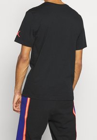 Černé bavlněné tričko s krátkým rukávem, kulatým výstřihem a červeným logem detailně umístěným na levém rameni.