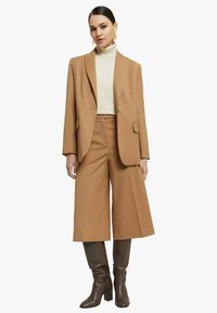 Completo con blazer color cammello e culottes, abbinato a un dolcevita crema. Presenta texture lisce e un design strutturato. Stivali marroni al ginocchio completano il look.