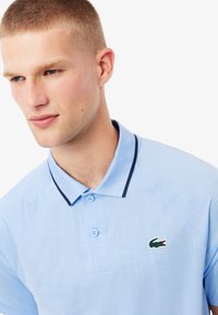 Ljusblå pikétröja med tvåknappsknäppning, marinblå kant på kragen och en grön krokodillogotyp på bröstet. Strukturerat tygmönster.