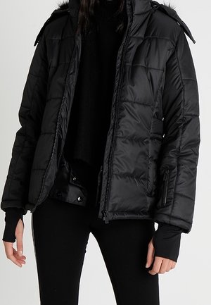 Femme portant une veste matelassée noire avec fermeture éclair et boutons-pression, associée à un pantalon noir et un haut à manches longues noir.