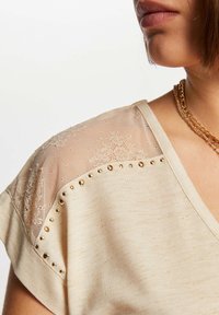 Top beige con una tela suave, con detalle de encaje en el hombro y acentos de tachuelas doradas a lo largo del escote.