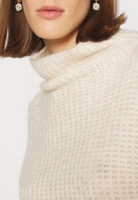 Pull en maille beige clair avec un col haut, présentant un motif texturé et des détails côtelés. Le modèle porte des boucles d'oreilles rondes en perles.