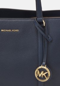 Sac à main en cuir bleu marine avec une texture granuleuse, équipé de quincaillerie dorée, d'un charme de logo et d'une forme structurée. Poignées pour le transport.