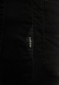 Jeans de mezclilla negra con un bolsillo diagonal distintivo y una etiqueta tejida que dice "CHASIN'". Textura suave y diseño ajustado.
