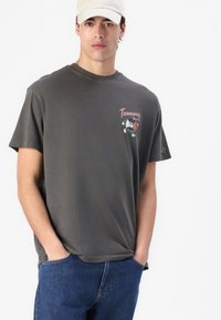 Šedé bavlněné tričko s kresleným grafickým motivem a textem "Tommy Jeans" na hrudi. Model k němu kombinuje modré džíny a světlou čepici.