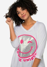 Weißes Langarmshirt mit V-Ausschnitt, das mit einem großen silberglänzenden Smiley-Gesicht und pinkem Text "DU BIST EINZIGARTIG" darunter verziert ist. Weicher Stoff.