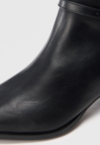 Černá kožená kotníková bota se hladkým povrchem a šitým páskem podél horního okraje, zobrazená na bílém pozadí.
