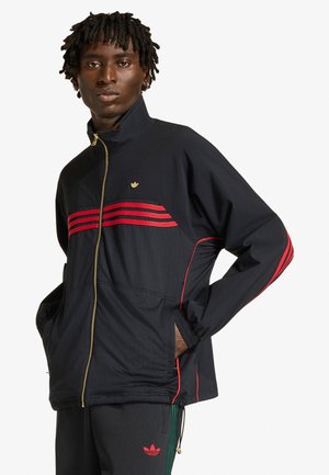 Giovane uomo con giacca Adidas nera con strisce rosse e cerniera dorata, mani nelle tasche, posa contro uno sfondo bianco semplice.
