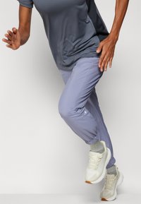 Lette, avsmalnede treningsbukser i lavendel. Glatt stoff med elastiske mansjetter, sammen med en mørkegrå treningsoverdeler og hvite joggesko.
