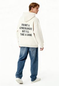 Felpa con cappuccio di colore chiaro con testo stampato nero sul retro. Abbinata a jeans blu larghi e sneakers bianche.