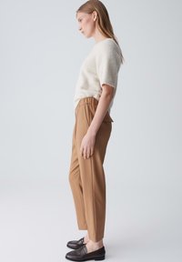 Hellbeige strukturierter Pullover mit kurzen Ärmeln, kombiniert mit beigefarbenen, schmal zulaufenden Hosen und dunklen Slippern, von der Seite vor einem einfarbigen Hintergrund betrachtet.